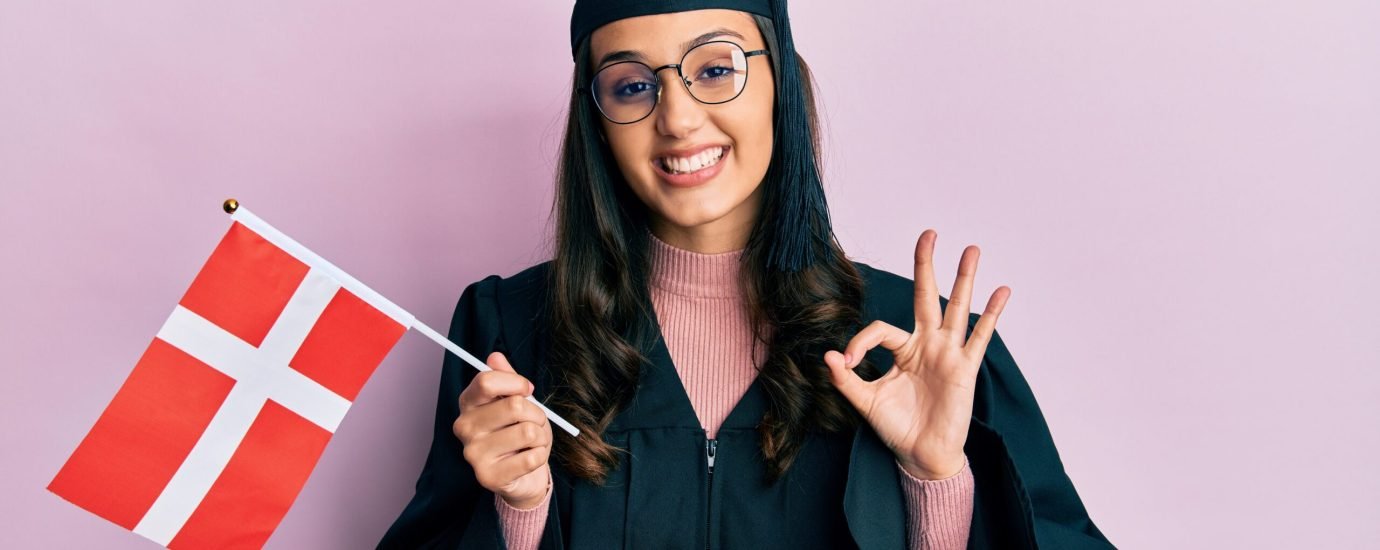 Young hispanic woman wearing graduation uniform holding denmark flag doing ok sign with fingers, smiling friendly gesturing excellent symbol Young hispanic woman wearing graduation uniform holding denmark flag doing ok sign with fingers, smiling friendly gesturing excellent symbol